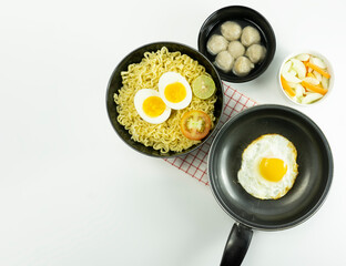 Indonesian Warm noodles served with eggs and fresh tomatoes with meatballs on a black bowl and pickled on a white bowl and eggs on a white background