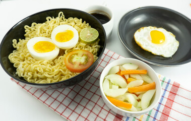 Indonesian Warm noodles served with eggs and fresh tomatoes with meatballs on a black bowl and pickled on a white bowl and eggs on a white background