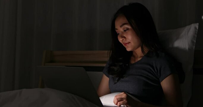Asian cute woman laying on bed at night in bedroom and using and typing on notebook computer laptop with happy face and comfortable gesture.
