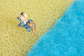 Miniature people toy figure photography. Men relaxing at beach chair in front of wavy beach