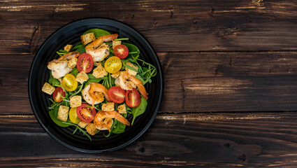 Top view flat lay shot of a homemade grilled shrimp salad with croutons, red and yellow cherry tomatoes in the black dish on the wooden table. Fresh prawn with vegetable. Concept of healthy food