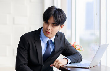 Asian young businessman are smart and handsome wearing black suit sitting while typing on keyboard at laptop near window in office at the morning