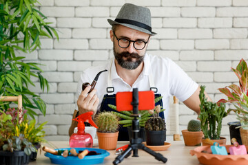 An internet seller, a beautiful beard man wearing hat and eyeglasses sitting in home indoor garden...