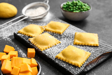 Wooden board with raw ravioli on dark background