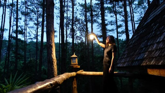 A young woman holding lantern and looking around the log cabin in the woods
