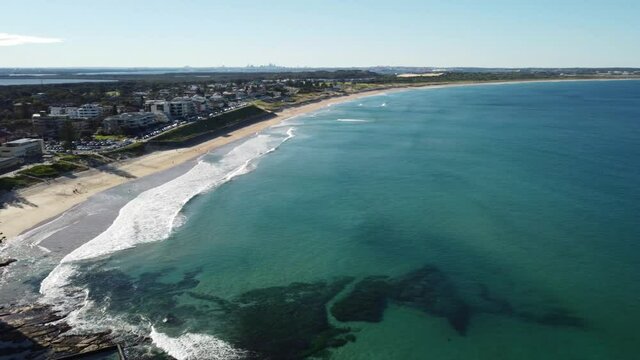 Beach and ocean with turquoise water, man made rock pool swimming pool, coastline, and apartments rising along the shore in aerial view under sunny sky