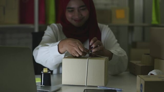 Slow motion of young muslim asian girl packing parcel using string scissors and tape on working desk with other boxes lying around. Deliver product to customers. Computer notebook in front.