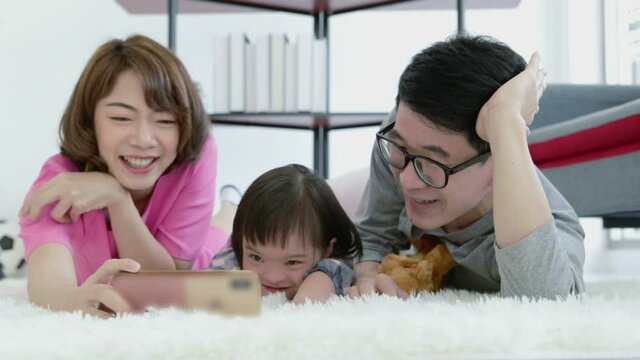 Slow motion of happy cheerful asian family of three father mother little daugther lying down on carpet watching cartoon with smartphone