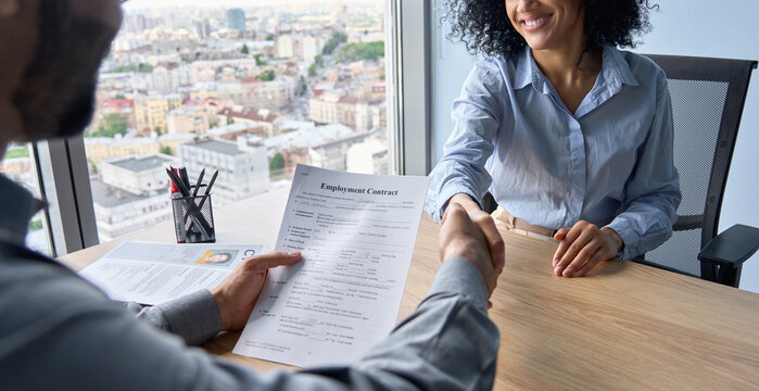 Closeup of Indian latin ceo holding employment contract job offer hiring welcoming female African American newcomer worker manager shaking hands in contemporary office. Human resources concept.