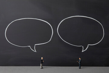 two miniature businessmen talking over chalkboard background with empty speech bubble