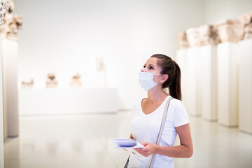 Portrait of young woman in protective face mask observing sculptures exposition in art museum,...