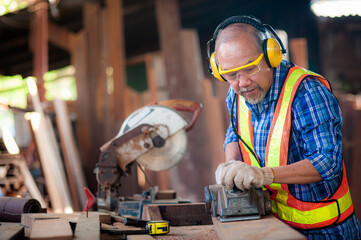 Asian elderly carpenter In work clothes with work safety equipment such as gloves,glasses, ear-muffs or noise-cancel equipment are using an electric planer to smooth out the wood to produce furniture.