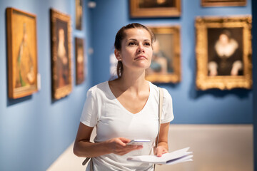 Focused adult girl holding brochure with exhibition program admiring paintings in museum