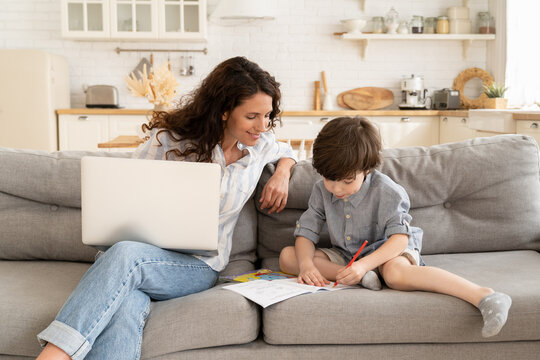 Home office and home education: young mother remote worker, freelance businesswoman sit with small preschool son on couch in living room working on laptop computer. Covid-19 lockdown lifestyle concept