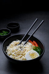 Black bowl with noodle, egg and vegetables and chopsticks near at black background.