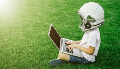 A child sits on the grass in an astronaut's helmet and studies on a laptop online enjoying nature..