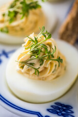 Kalte gefüllte russische Eier mit Frischkäse, Kresse und Kräuter farciert und Toast Brot auf Blau Weiß Porzellan Eierplatte und Marmor Hintergrund hell in Detail Nahaufnahme 