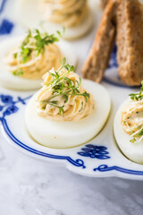 Kalte gefüllte russische Eier mit Frischkäse, Kresse und Kräuter farciert und Toast Brot auf Blau Weiß Porzellan Eierplatte und Marmor Hintergrund hell in Detail Nahaufnahme 