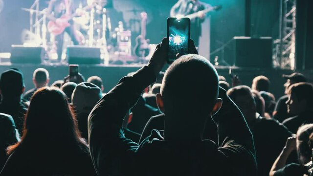 People filming rock concert on smartphones. Silhouettes crowd of people dancing at an open-air music festival. Fans applaud, raise hands up, and photographing. Party People in Action. 4K, Slow Motion