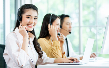 Focus Asian beautiful attractive female call center cheerful smiling with headsets, sitting, looking camera, monitoring with colleagues in office or agency at computer, giving service to customers.