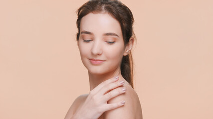 Young brown-haired beauty model gently touches her shoulder, sliding her hand down her arm, turns and looks at the camera and smiles | Posing for skin care product commercial.