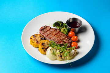 Delicious grilled beef steak with bbq vegetables and corn served on a white plate with sauce over blue background.