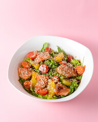 Fresh salad with salmon and vegetables close up in a bowl over pastel pink background.