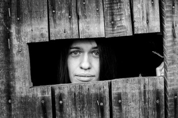 Young woman looking through the crack of a locked wooden shed, close-up. Black and white photo.