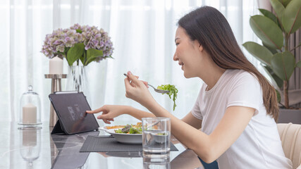 Asian woman eating fresh salad and using tablet at her home dining table, Vegetable salads are rich in vitamins and minerals, Fat-low-calorie and high-fiber diets, Healthy food, Appetizer.