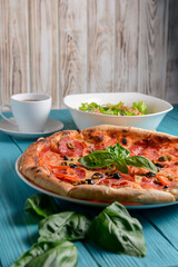 Fresh italian pizza with salami and black olives and other food on the background. Blue colored wooden table.