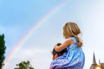 Little preschool girl sitting on shoulder of father. Happy toddler child and man observing rainbow on sky after summer rain. Happy family, bonding, love. Summertime. Dad and daughter. Father's day.