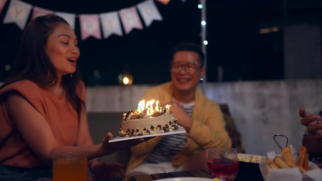 Diversity Asian millennial people friends enjoy celebration birthday party together at outdoor rooftop with food and drink. Woman excited with birthday cake and blowing birthday candle with happiness