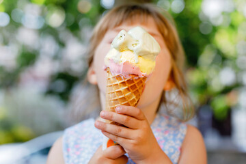 Little preschool girl eating ice cream in waffle cone on sunny summer day. Happy toddler child eat...