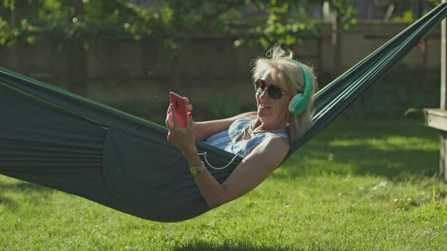 caucasian senior lady swings in the hammock listening to music with phone and headphones.