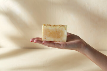 natural cosmetics, hygiene and beauty concept - hand holding bar of craft soap on beige background