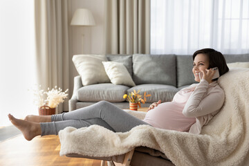 Fototapeta na wymiar Full length happy pregnant woman chatting on phone at home, barefoot attractive happy young future mom relaxing in cozy chair, holding smartphone and talking, enjoying pleasant conversation