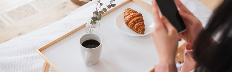 cropped view of young brunette woman holding cellphone near table with coffee and croissant in bedroom, banner