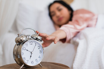 sleepy young asian woman touching alarm clock on bedside table in bedroom