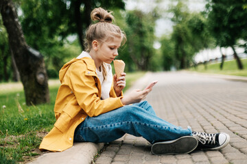 A blond girl sitting on the road with a phone and ice cream communicates in social networks. Child outdoors
