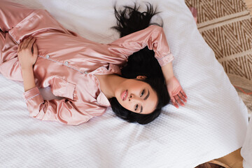 top view of young asian woman in silk pajamas lying on bed with white linen in bedroom