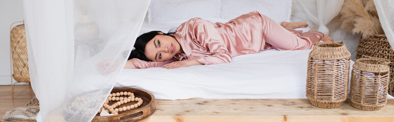 young asian woman in silk pajamas lying with outstretched hand on white linen in bedroom, banner