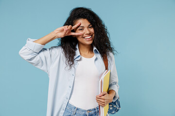 Young african american girl teen student wear denim clothes backpack hold books cover eye with victory gesture isolated on blue background studio. Education in high school university college concept