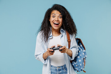 Young excited fun african american girl teen student wearing denim clothes backpack play pc game with joystick console isolated on blue background Education in high school university college concept