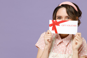 Fun cheerful happy young housewife housekeeper chef cook baker woman in pink apron hiding cover mouth with gift voucher flyer mock up isolated on pastel violet background studio Cooking food concept.