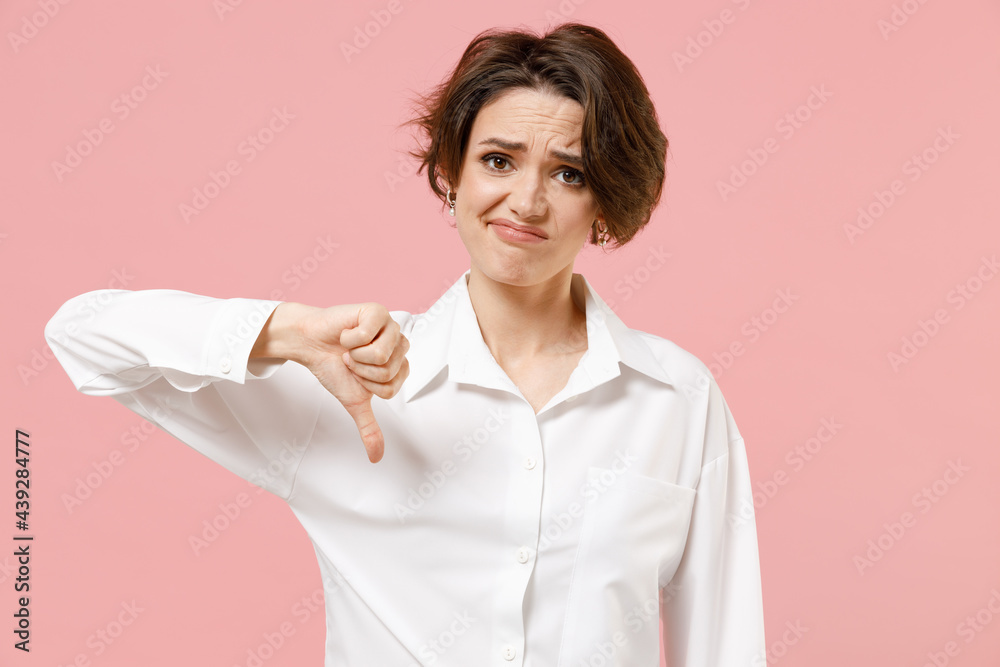 Wall mural Young displeased dissatisfied employee business woman corporate lawyer in classic formal white shirt work in office showing thumb down dislike gesture isolated on pastel pink color background studio.