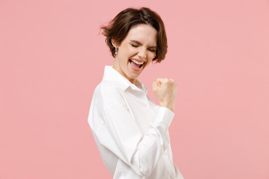 Young fun overjoyed successful employee business woman corporate lawyer in classic formal white shirt work in office do winner gesture clench fist say yes isolated on pastel pink background studio.