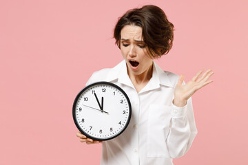 Young sad displeased employee business secretary woman corporate lawyer in classic formal white shirt work in office hold clock spread hands isolated on pastel pink color background studio portrait.