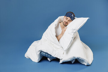 Full size body length young man 20s in pajamas jam sleep mask resting relax at home sleep on pillow sit wrap cover with blanket duvet isolated on dark blue background Good mood night bedtime concept.