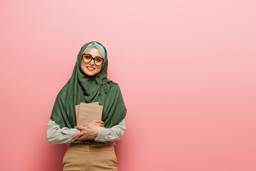 Fototapeta premium pretty muslim teacher with textbooks smiling at camera on pink background