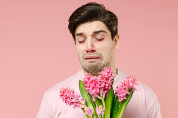Sick ill allergic man has red eyes runny stuffy sore nose suffer from pollen allergy symptoms hay fever hold blooming flower plant reaction on trigger isolated on pastel pink color background studio.
